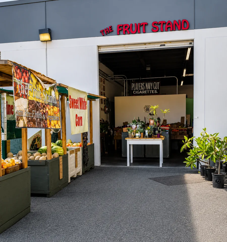 The entrance of The Fruit Stand with farm-fresh produce banners, sweet white corn and melons out front