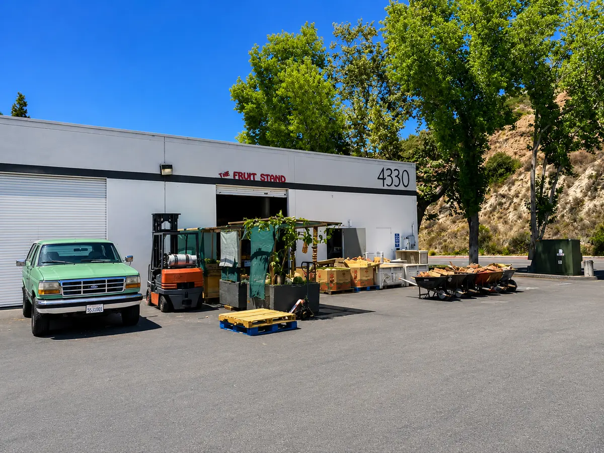 The exterior of The Fruit Stand at 4330 Morena Blvd: a warehouse bay with a vintage green pickup, produce stand and firewood, San Diego hills behind
